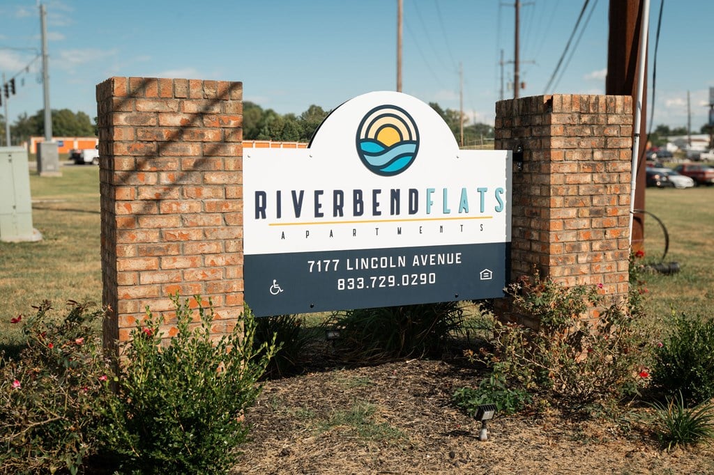 A sign for Riverbend Flats is displayed in front of a brick wall.