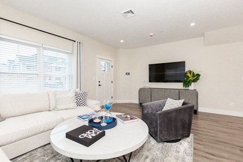 A living room with a white couch, a grey armchair, a coffee table with a black book on it, and a flat screen TV mounted on the wall.