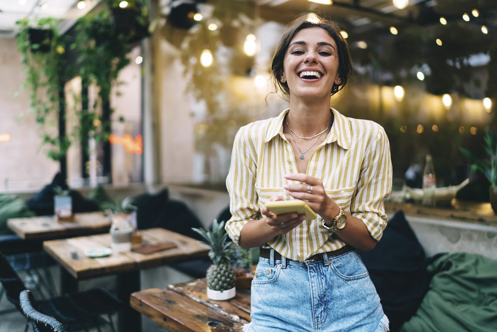 A woman in a striped shirt and jeans is laughing while holding a phone in a cozy, dimly lit room with hanging plants and tables at Mayla Pompano, Pompano Beach, FL