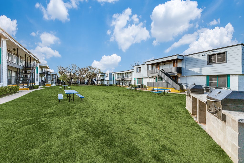 A grassy area in front of apartment buildings with picnic tables.