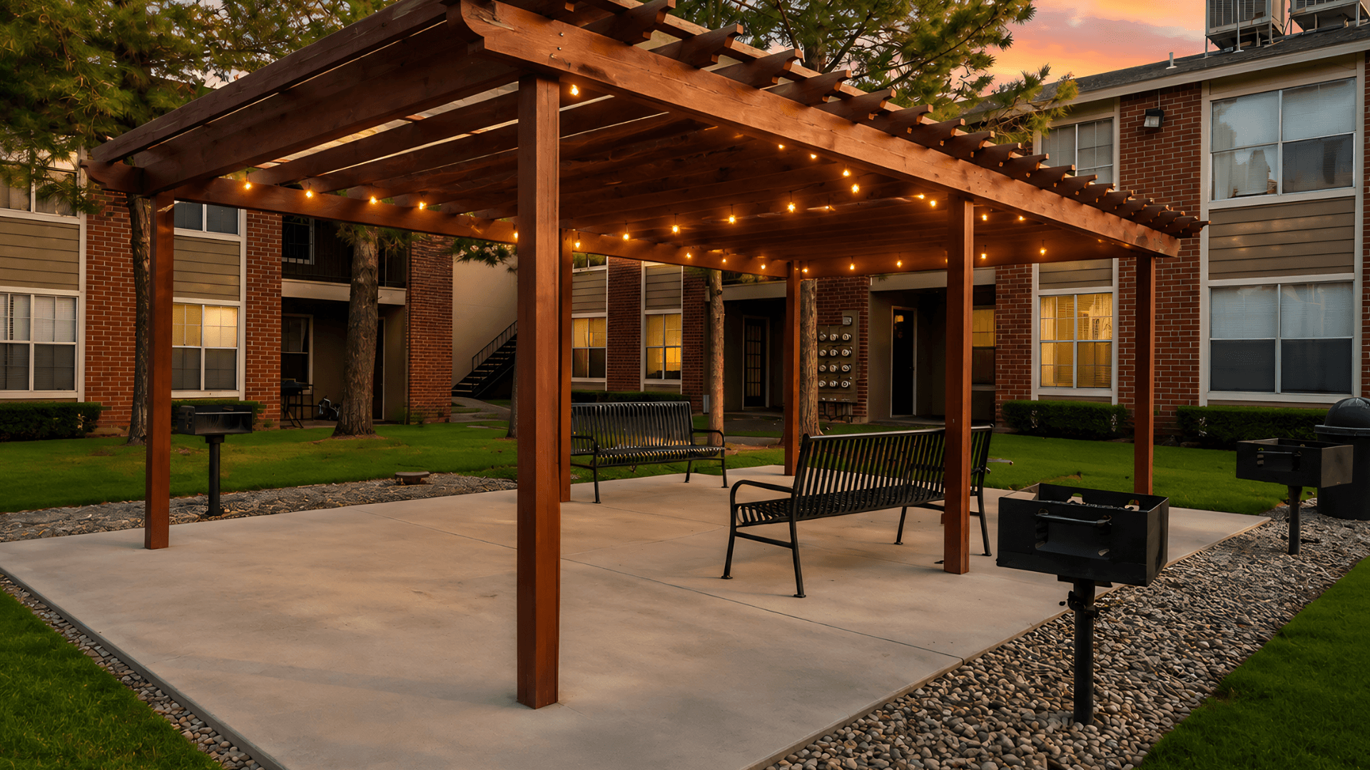 A wooden pergola with string lights is situated in a courtyard with benches and a mailbox.