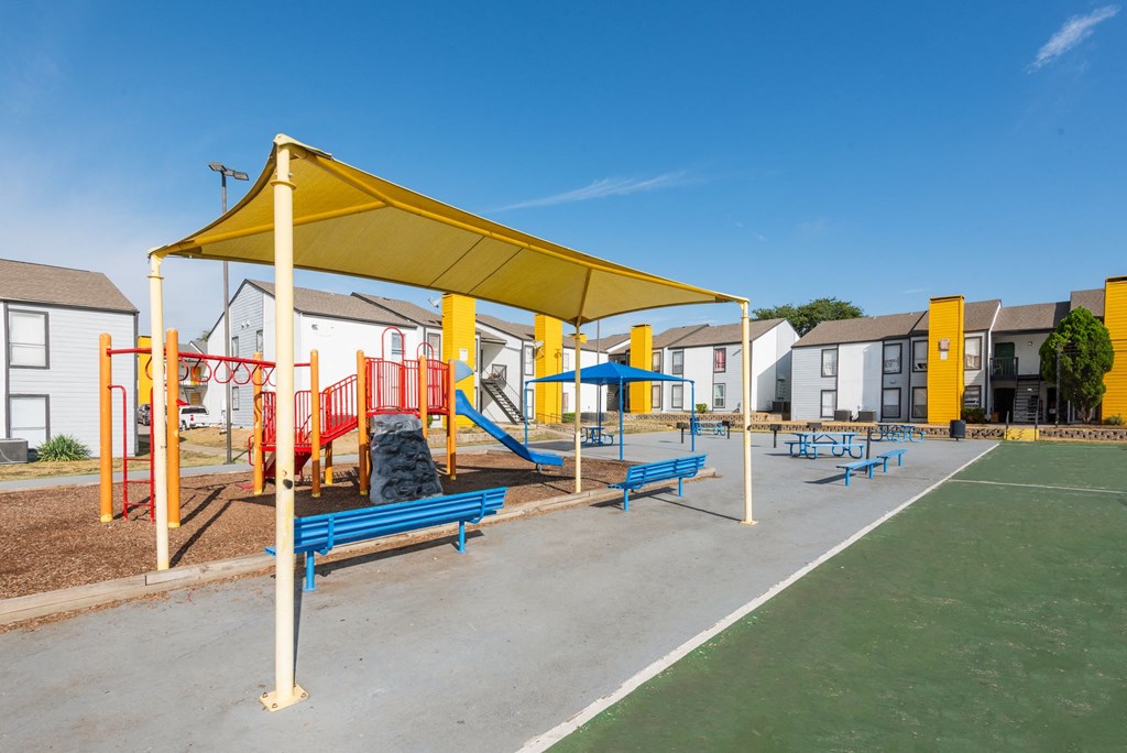 a playground with benches and a yellow canopy in front of apartment buildings at Helios, Dallas, TX