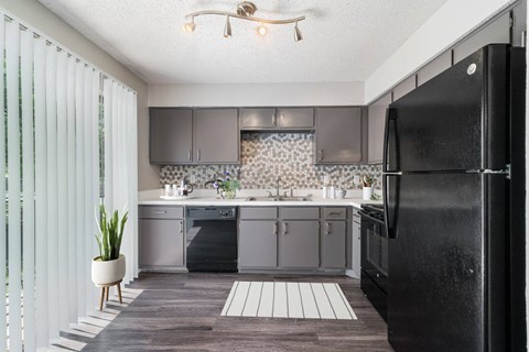 A black refrigerator is in the corner of a kitchen with wooden floors and white walls.