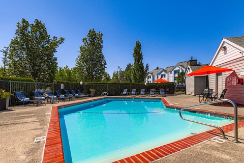 Happy Valley, OR, Apartments - Sunnyside Village - an Outdoor Gated Pool Area with Lounge Seating, Tables with Chairs and Umbrellas, Surrounding Trees and Shrubbery, and a View of the Property Building