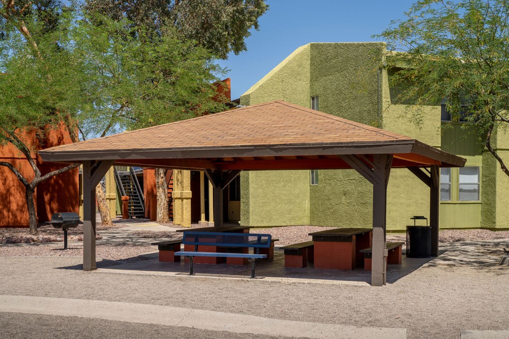 A pavilion with a brown roof is surrounded by a gravel area and a green building in the background.