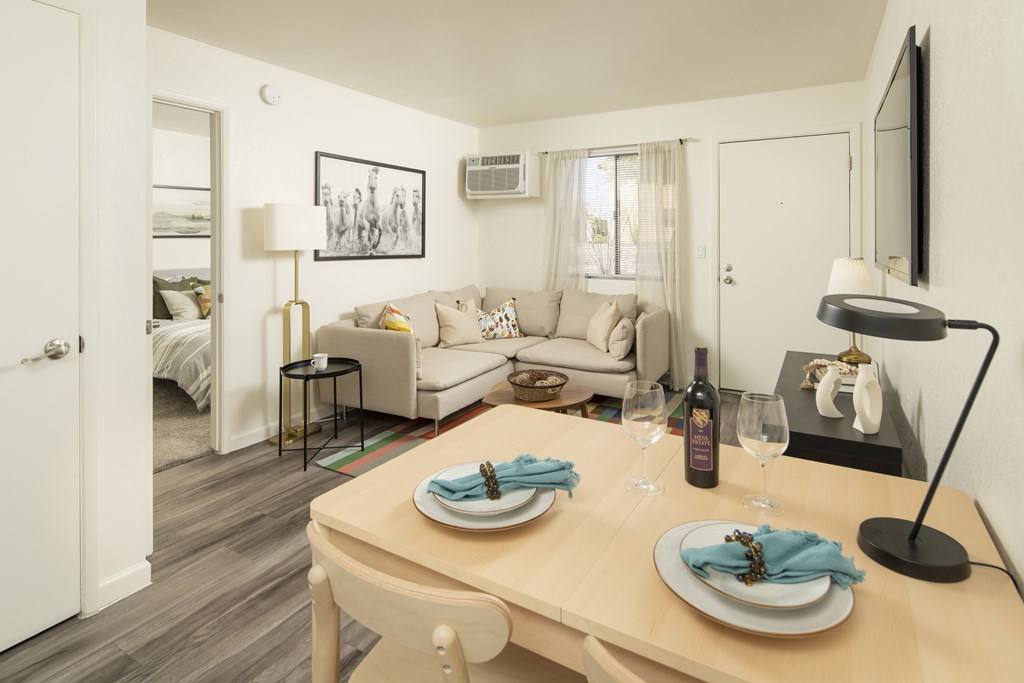 A dining table set with plates and glasses is in the foreground of a well-lit room with a couch and a bed in the background.