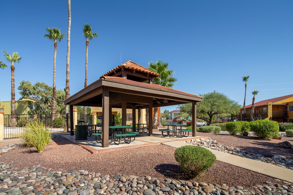 A gazebo with a red roof is surrounded by palm trees and a gravel area with a bench.