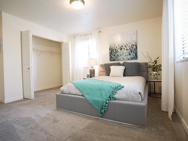 A bedroom with a bed, a grey headboard, a white bedspread, a green throw, a white pillow, a grey armchair, a white wall, a white door, a white curtain, a white lamp,