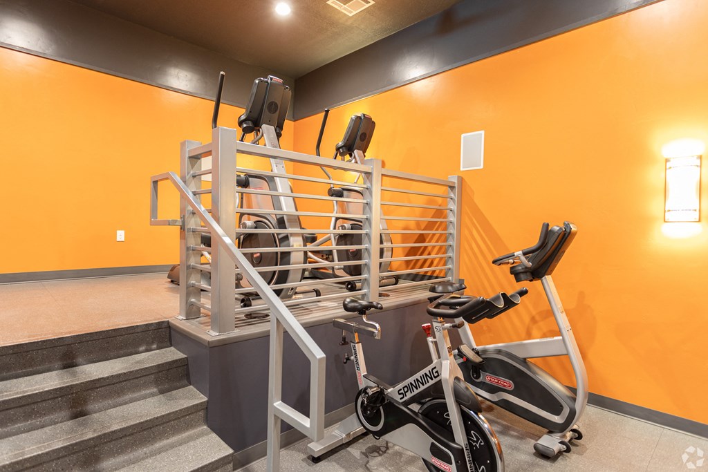 a gym with exercise equipment, ellipticals, and stairs in a building with orange walls at Outlook Apartments for rent in Springville and Provo, UT.