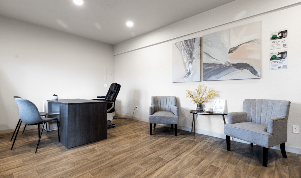 A room with a grey sofa, two chairs and a table with a vase of flowers on it.