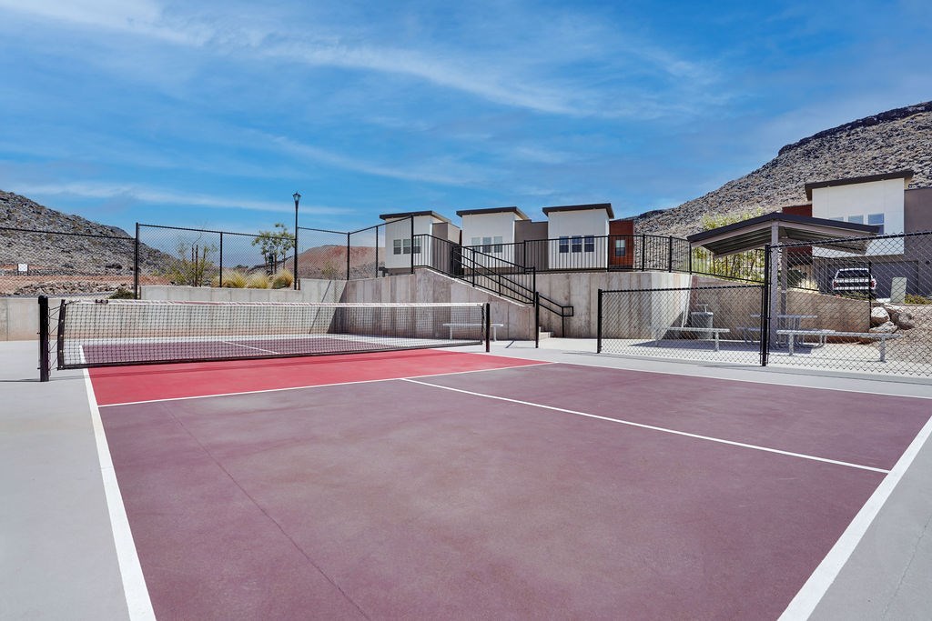 A tennis court with a red surface and white lines, surrounded by a fence and buildings in the background.