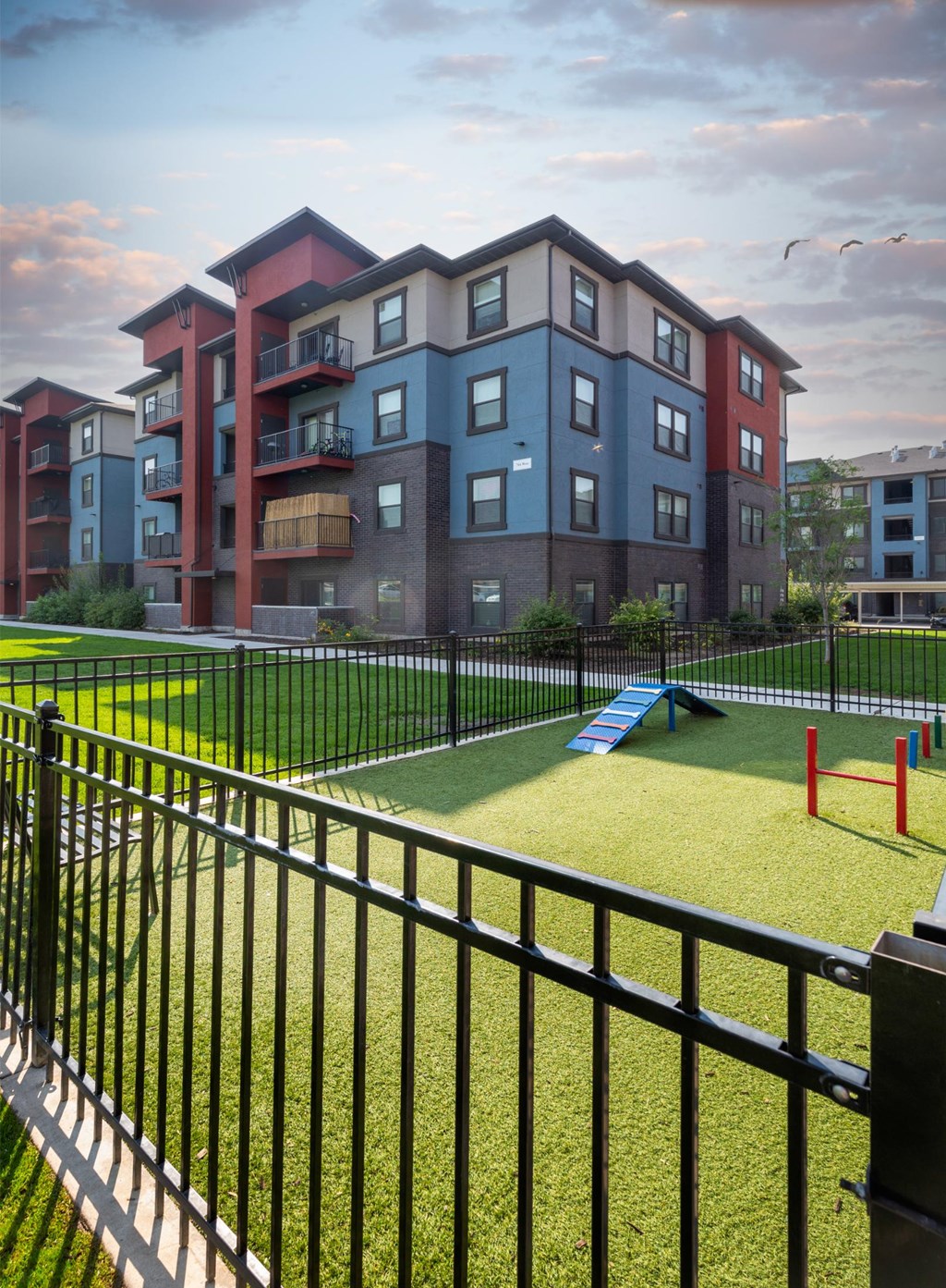 A black fence surrounds a playground in front of a multi-story apartment building.