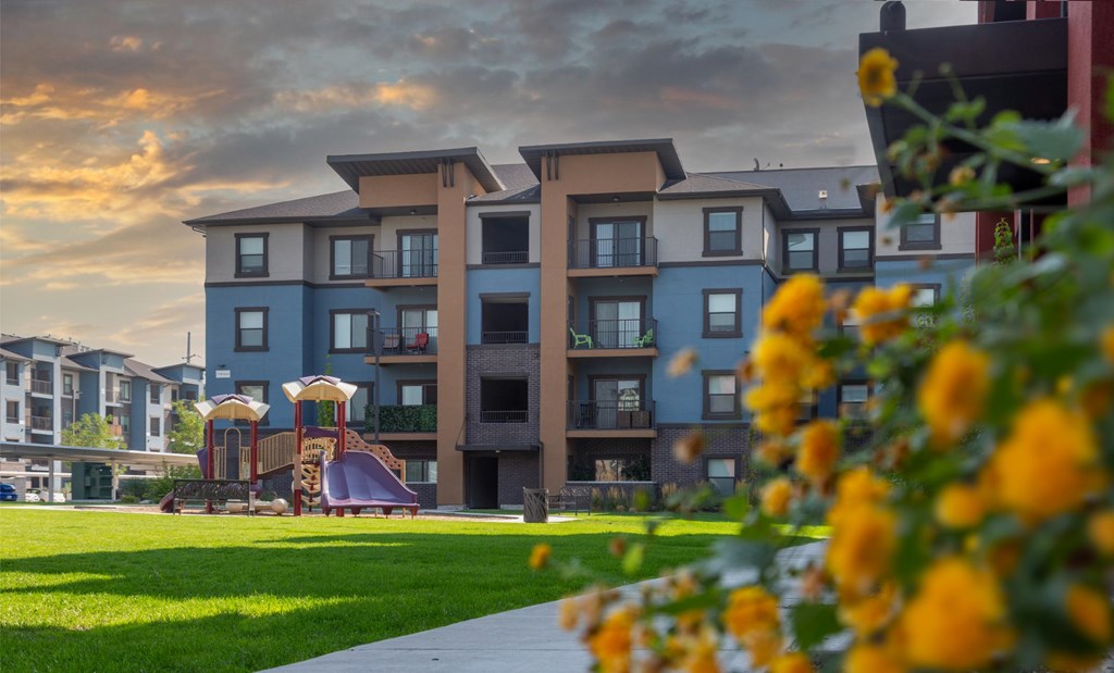 A modern apartment complex with a playground and flowers in the foreground.