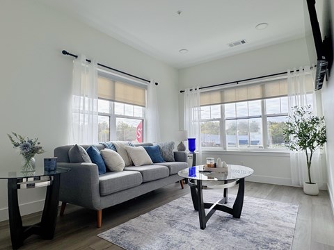 A living room with a grey couch, a black coffee table, and a large window with white curtains.