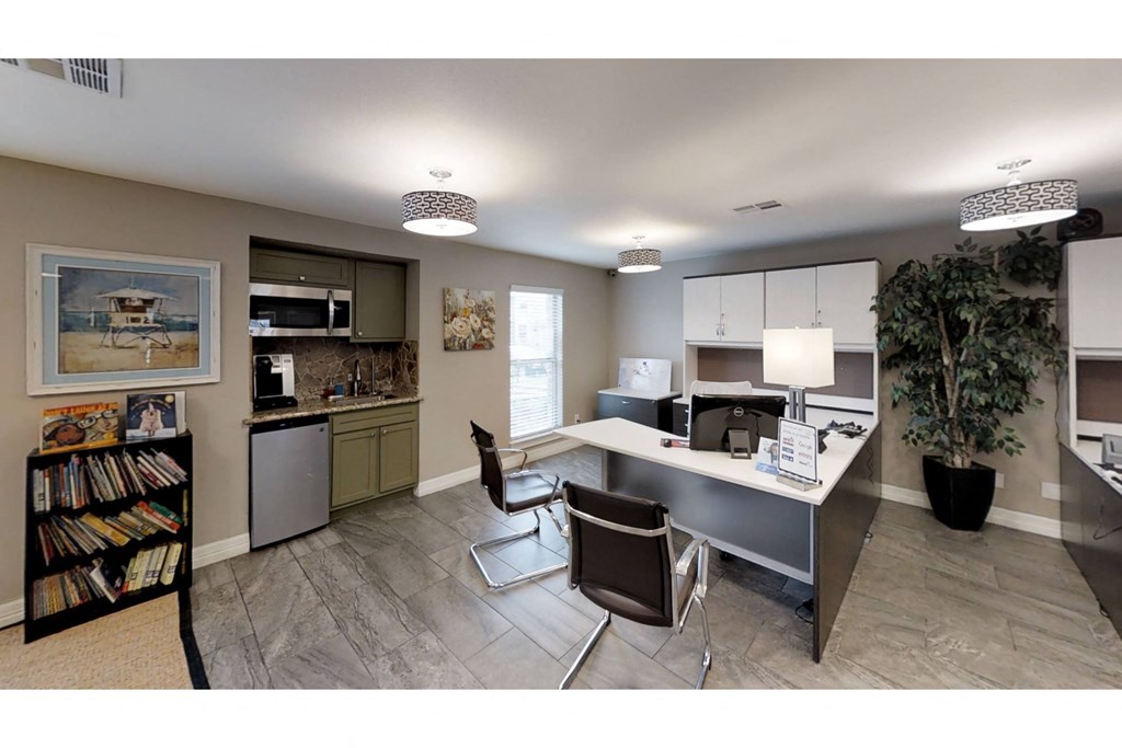 a room with a desk and chairs and a kitchen in the background