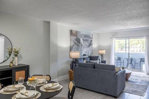 A modern dining room with a grey couch, a round table set for two, and a view of the balcony.