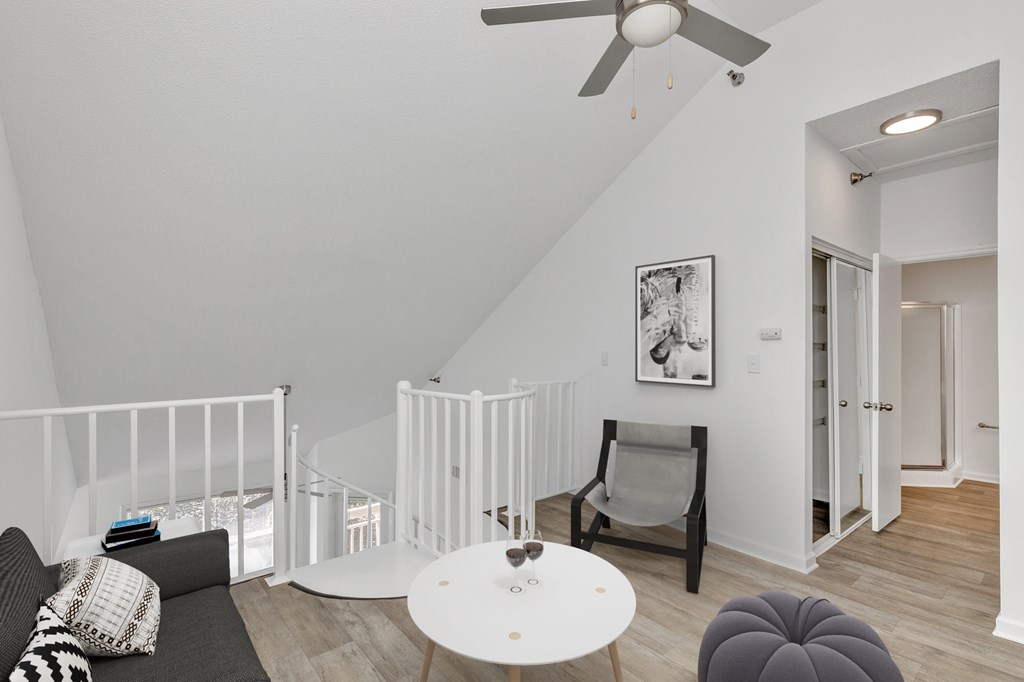 A living room with a grey couch, a white table, a grey ottoman, a white chair, a white railing, a white ceiling fan, and a white wall with a picture hanging on it.