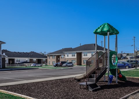 A playground with a green umbrella and slide in front of apartment buildings.
