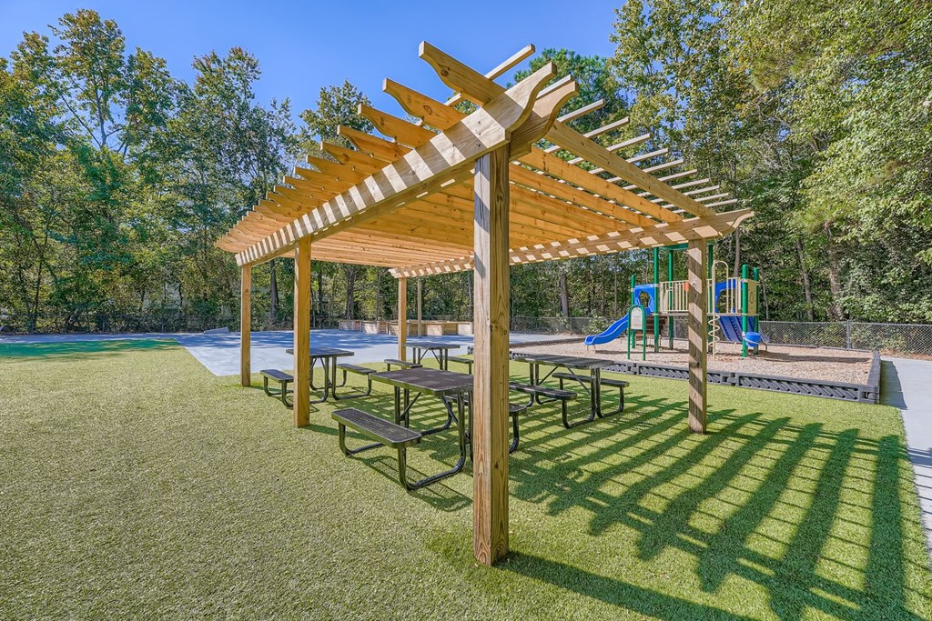 A wooden pergola with benches is situated in a grassy area with a playground in the background.