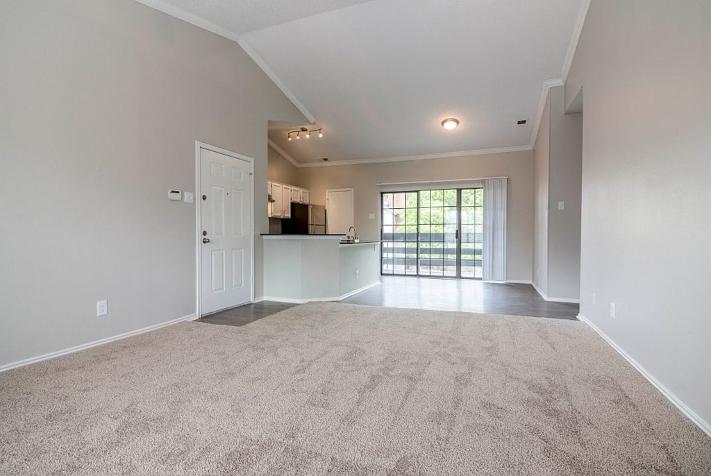 A spacious living room with a carpeted floor and a sliding glass door leading to a balcony.