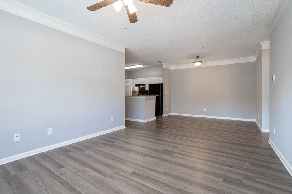 an empty living room with a ceiling fan and a kitchen in the background