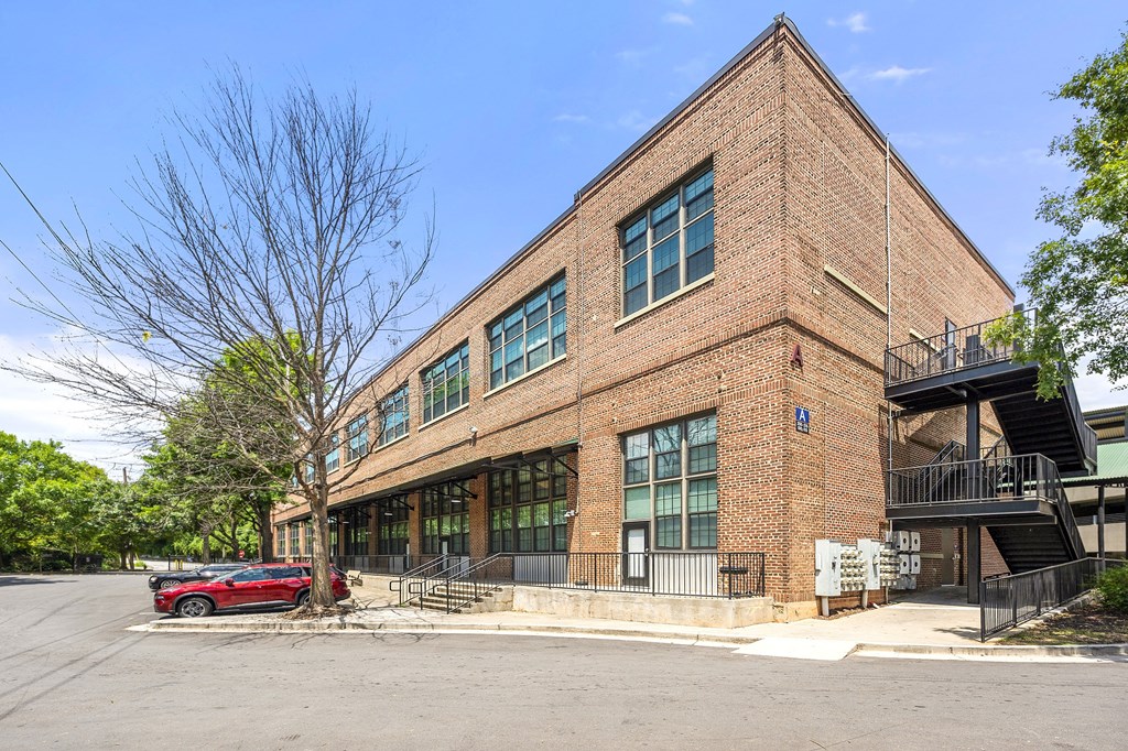 A red car is parked in front of a brick building with a tree in front of it.