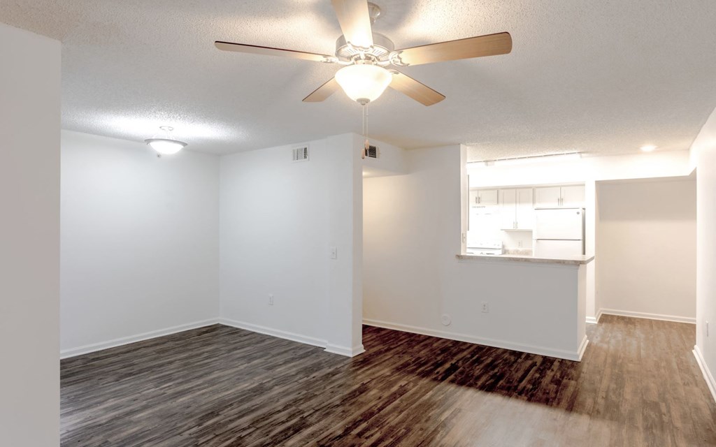 an empty living room with a ceiling fan and a kitchen in the background