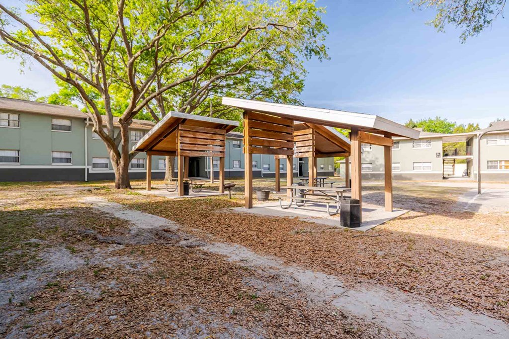 A wooden pavilion with a white roof is surrounded by a gravel area and a tree with green leaves.