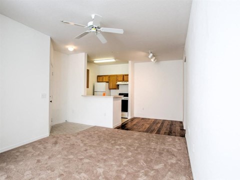 an empty living room with a ceiling fan and a kitchen in the background