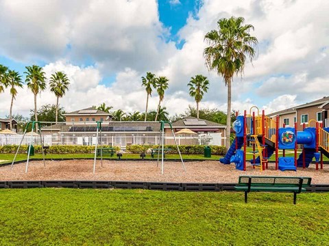 A playground with a slide, swings, and a bench is surrounded by palm trees.