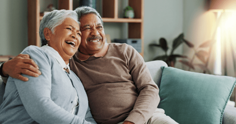 An older couple is sitting on a couch, smiling and laughing together.