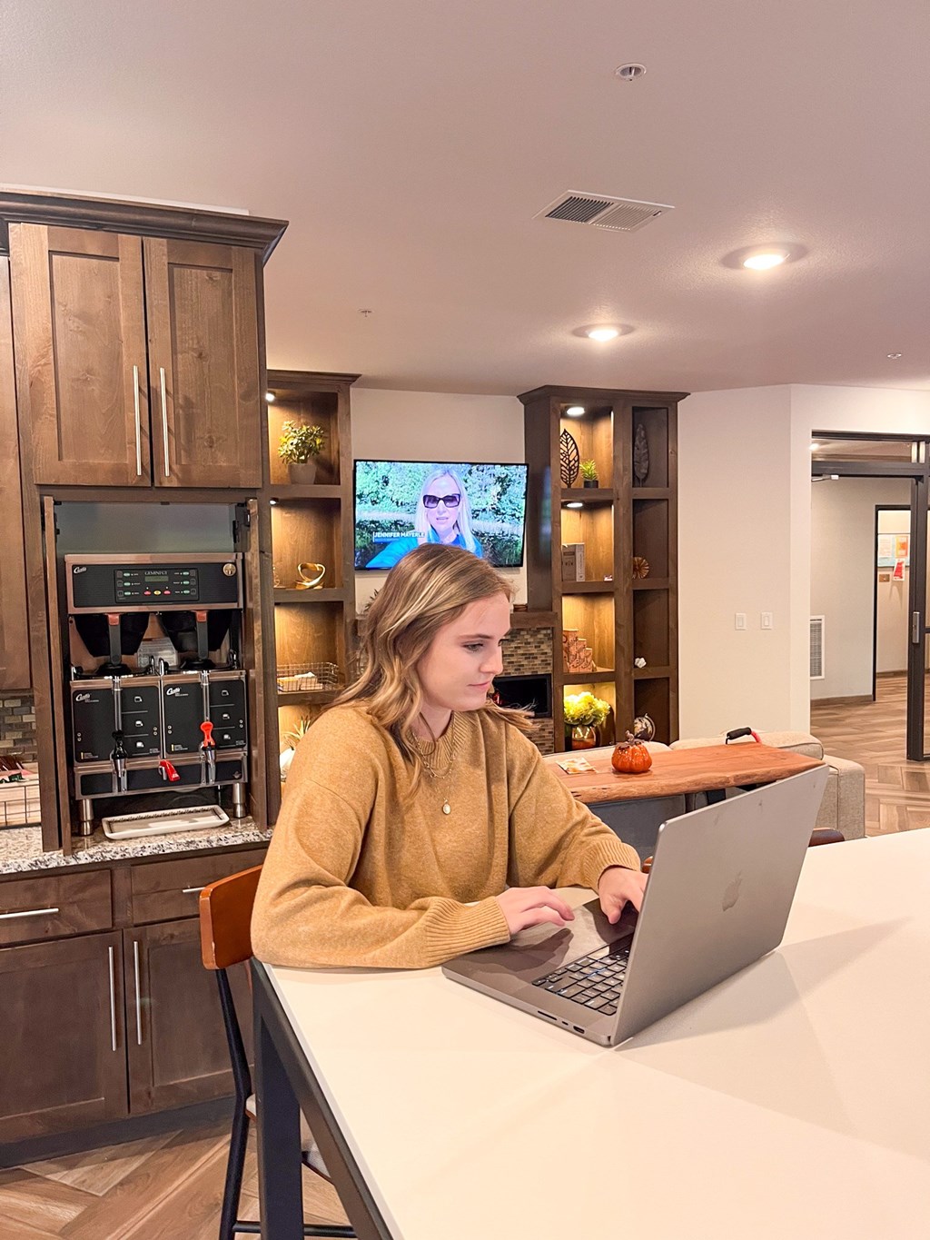 A woman in a yellow sweater is working on a laptop in a modern kitchen.