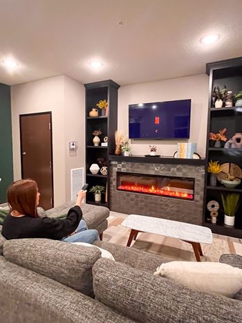 A woman is sitting on a grey couch in front of a fireplace and a television.