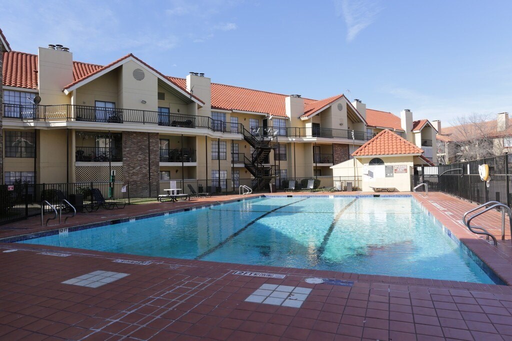A large outdoor swimming pool surrounded by a red tile floor and apartment buildings.