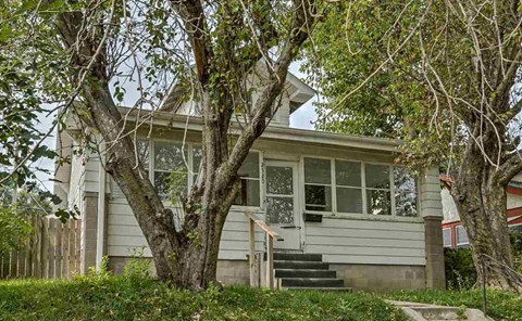 A house with a grey facade and a white door is surrounded by trees.