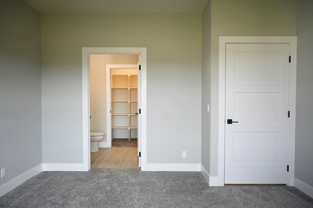 A room with a white door and a hallway with a toilet and a shelf.