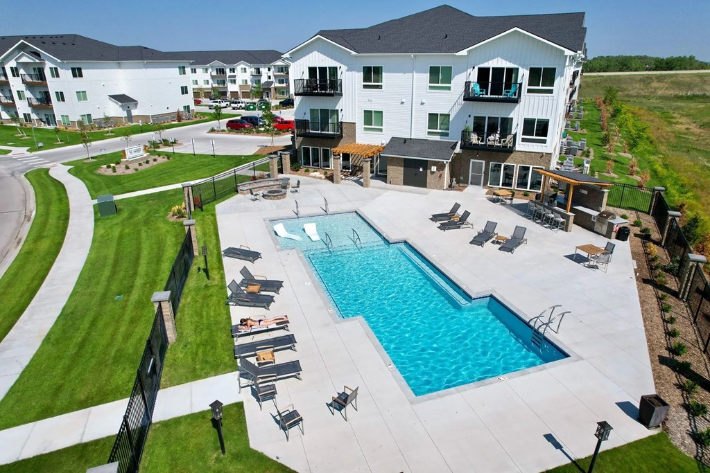 A large swimming pool surrounded by lounge chairs and apartment buildings in the background.
