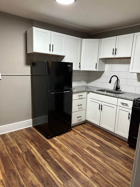 A black refrigerator in a kitchen with white cabinets and a wooden floor.