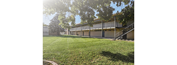 Exterior view of a single story apartment building with covered walkway, mature trees and green lawn at Kearney Apartment Homes.