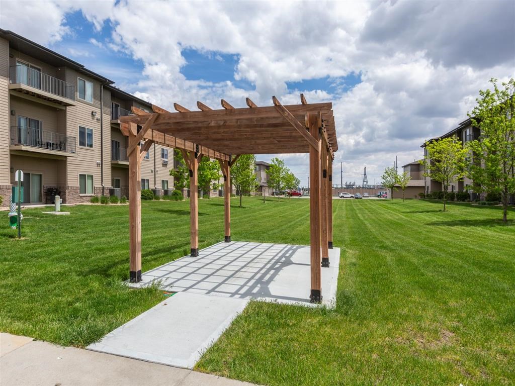 A wooden pergola structure is situated in a grassy area in front of apartment buildings.