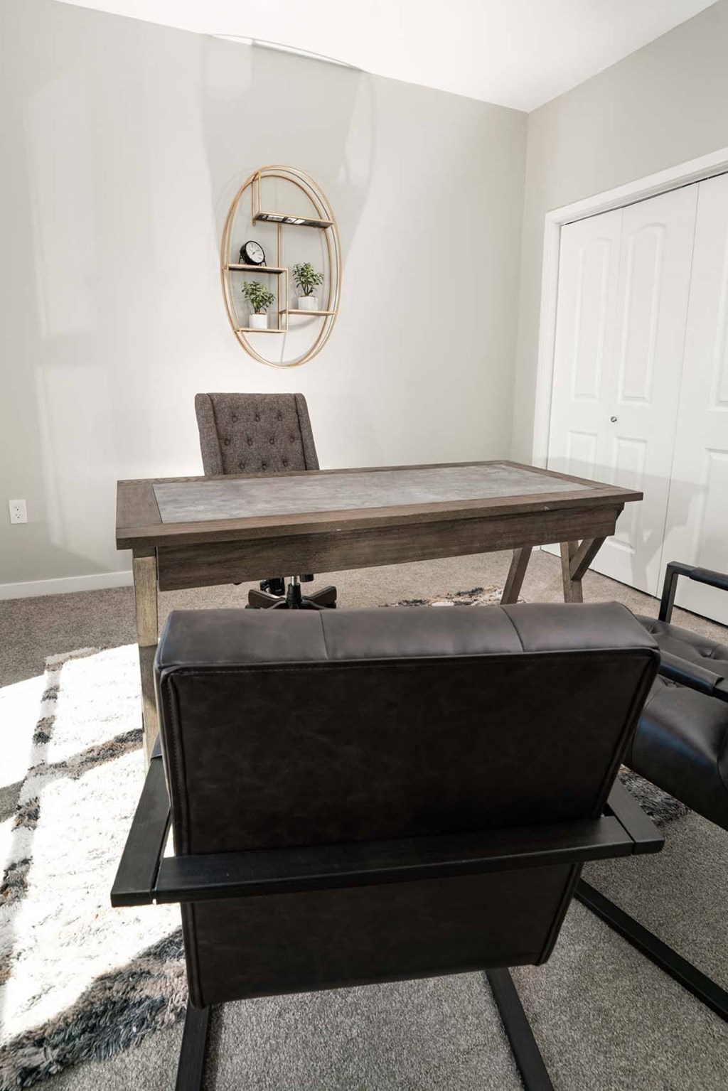 A brown leather ottoman sits in front of a wooden desk with a chair and a round mirror on the wall.