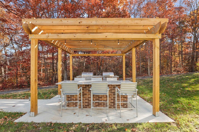 A wooden pergola with a grill and seating area in the middle of a grassy area.