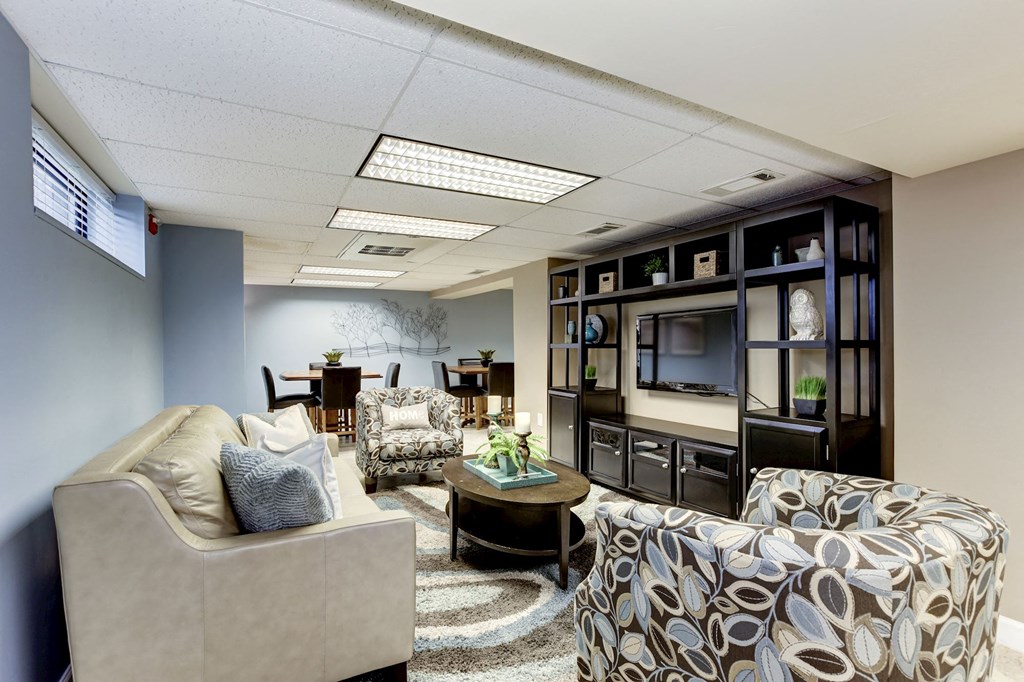 A living room with a beige couch, a patterned ottoman, and a bookshelf filled with decorative items.