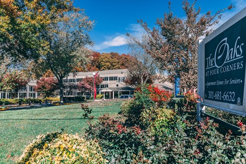 A sign for The Oaks at Four Corners is in the foreground of a grassy area with trees.