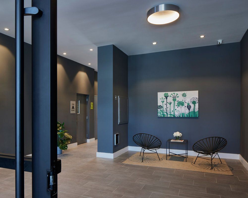 A hallway with a painting on the wall and two chairs with a table in front of them.