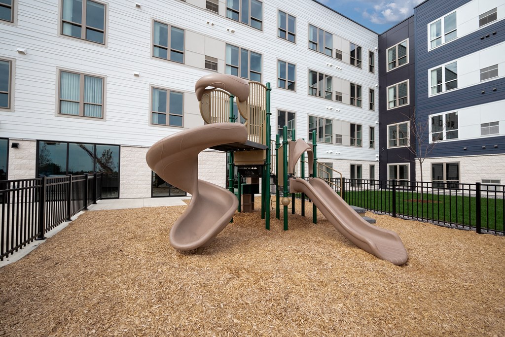 a playground with two slides and a chair in front of an apartment building at The Landing at Amber Fields Apartments, Rosemount, Minnesota