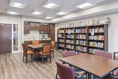 a library with tables and chairs and a kitchen and library shelves at The Winslow 62+ Apartments, West St Paul, Minnesota