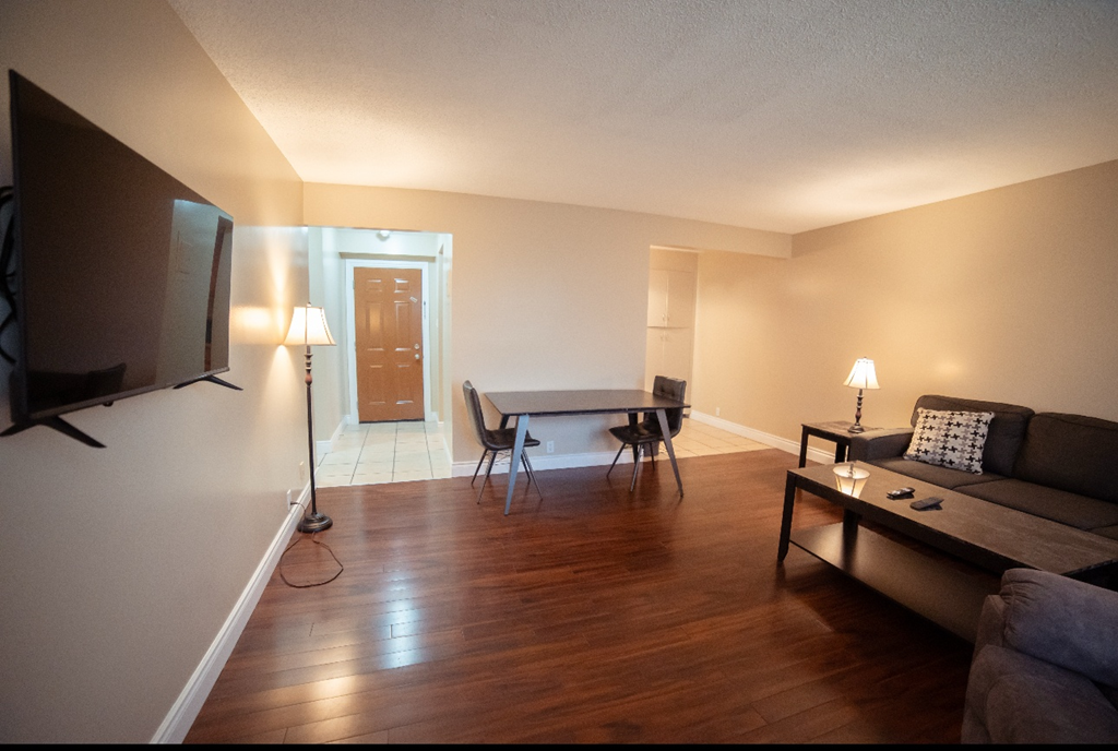 A living room with a grey couch, a black chair, a black table, a white tablecloth, a white lamp, a white door, and a white wall.