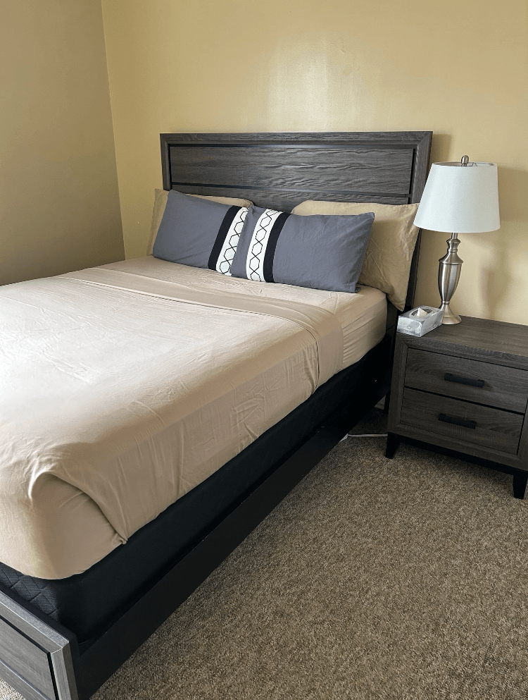 A bed with a white sheet and two pillows with a grey headboard and a nightstand with a lamp on it.