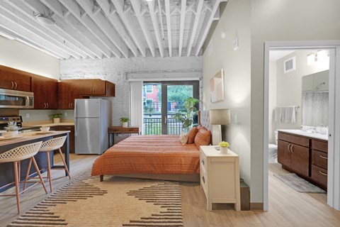 A bedroom with a bed, nightstand, and a kitchen area with a refrigerator and microwave.